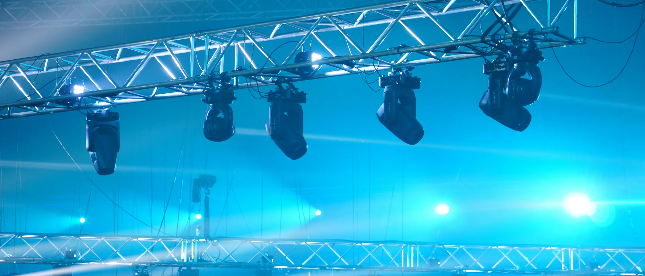Lighting truss with moving fixtures cutting blue beams through theatrical haze.
