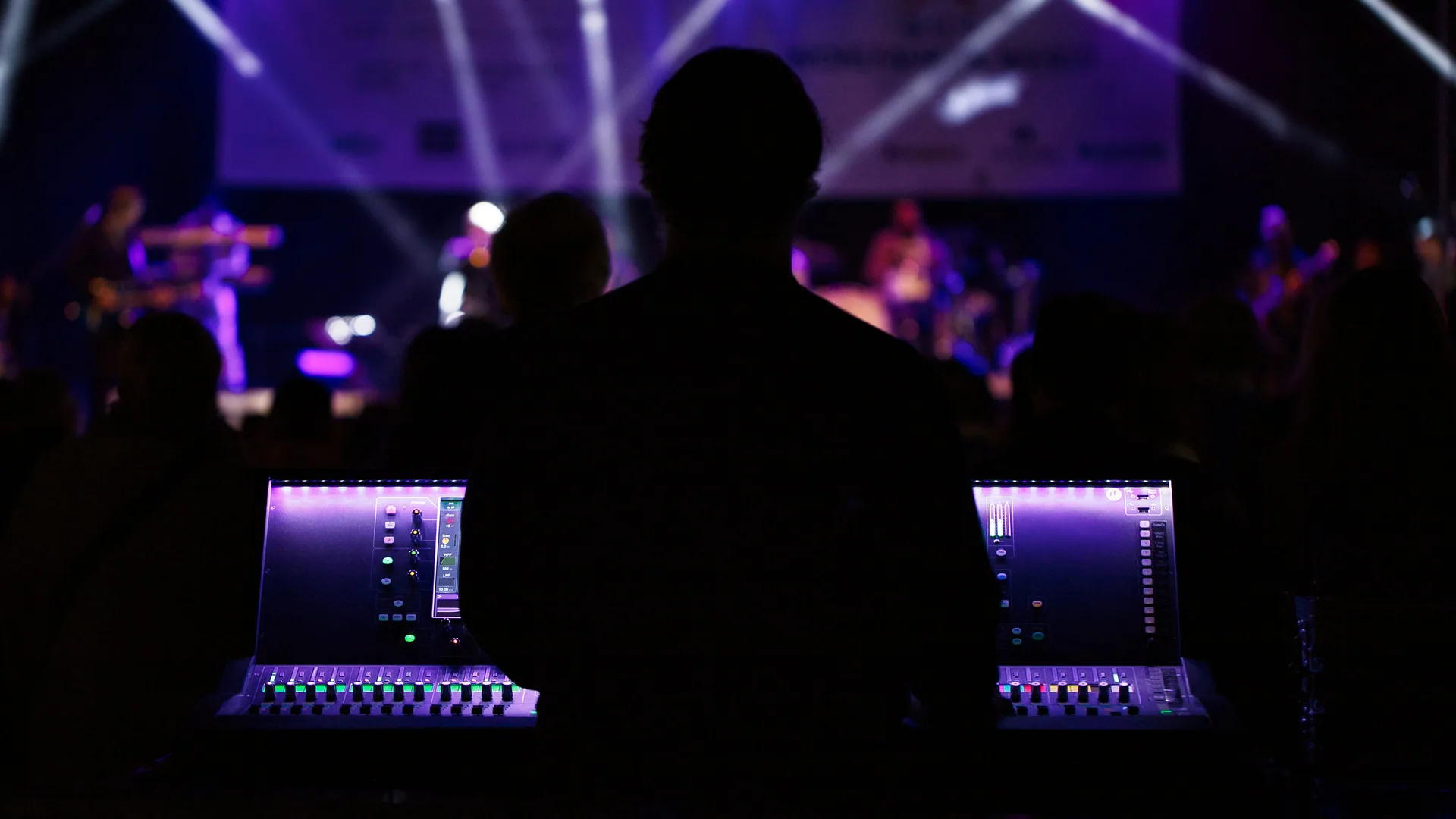 Front-of-house consoles illuminated under stage lights, engineer silhouetted at the desk.